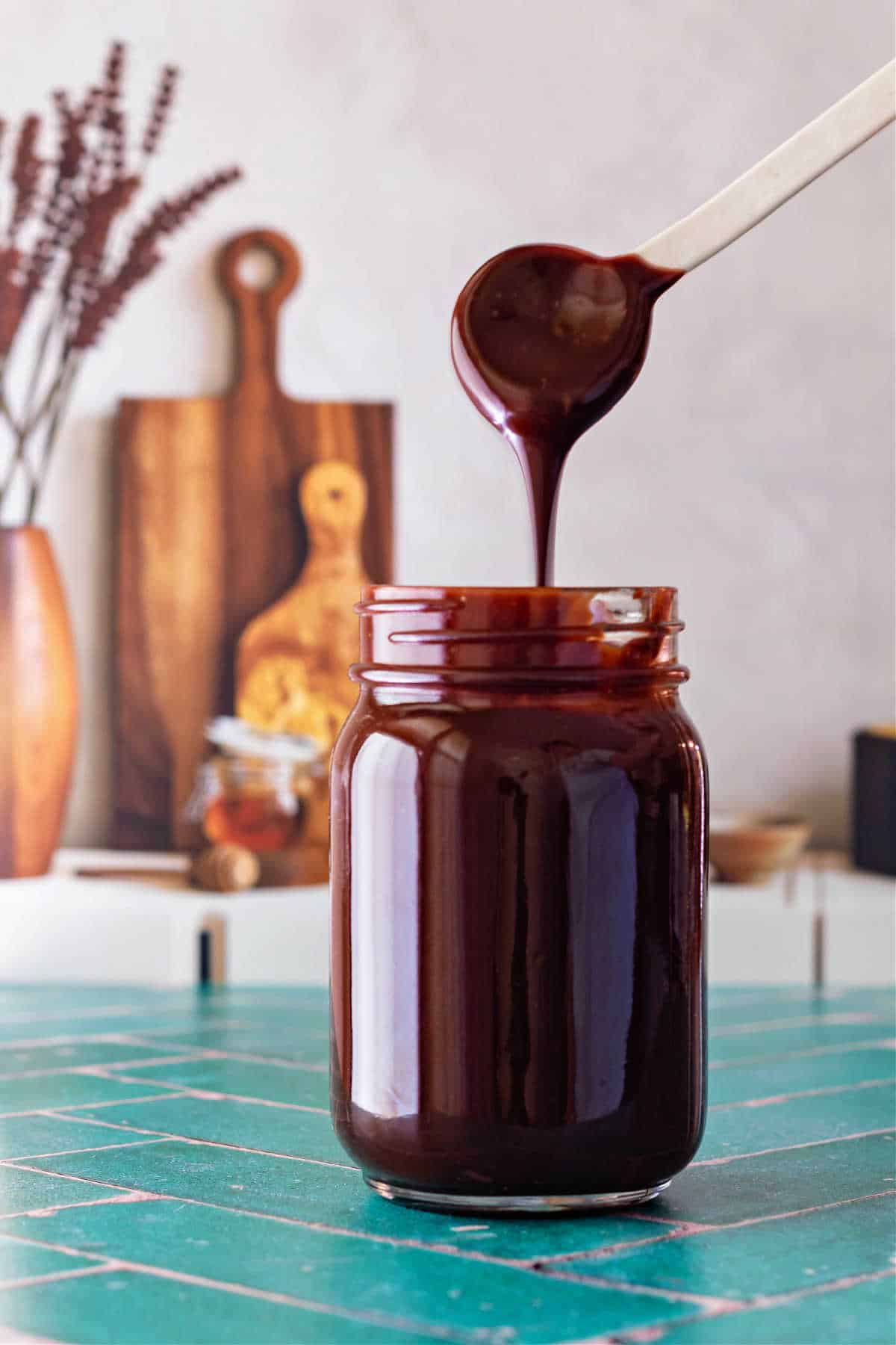 A white spoon with hot fudge sauce drizzling off it and back into the jar below.