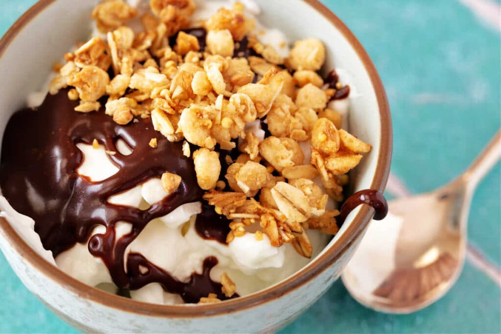A close-up image looking into a pale blue bowl with vanilla yogurt, hot fudge sauce, and granola in it.