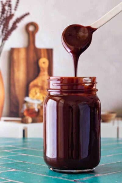 A white spoon with hot fudge sauce drizzling off it and back into the jar below.
