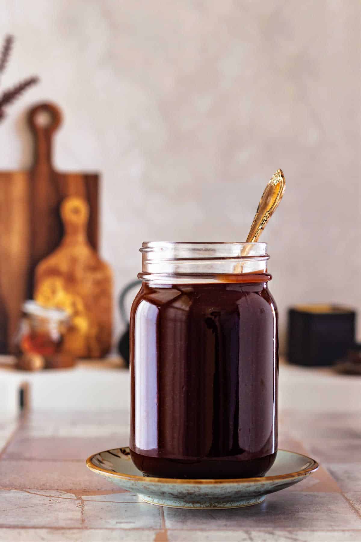 A glass jar full of fudge sauce with a spoon in it. It's shot on small gray tiles with a couple of cutting boards propped up in the background.
