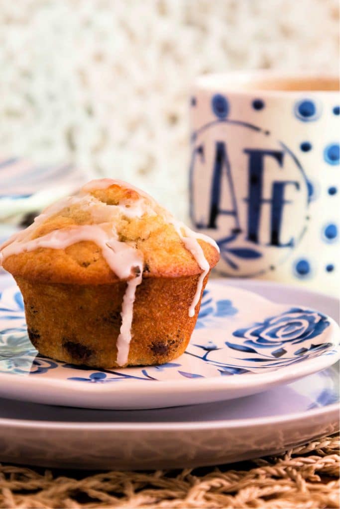 A glazed chocolate chip muffin on a plate with a cup of coffee ready for serving.