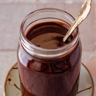 A square image looking down into a jar of fudge sauce with a spoon in it.