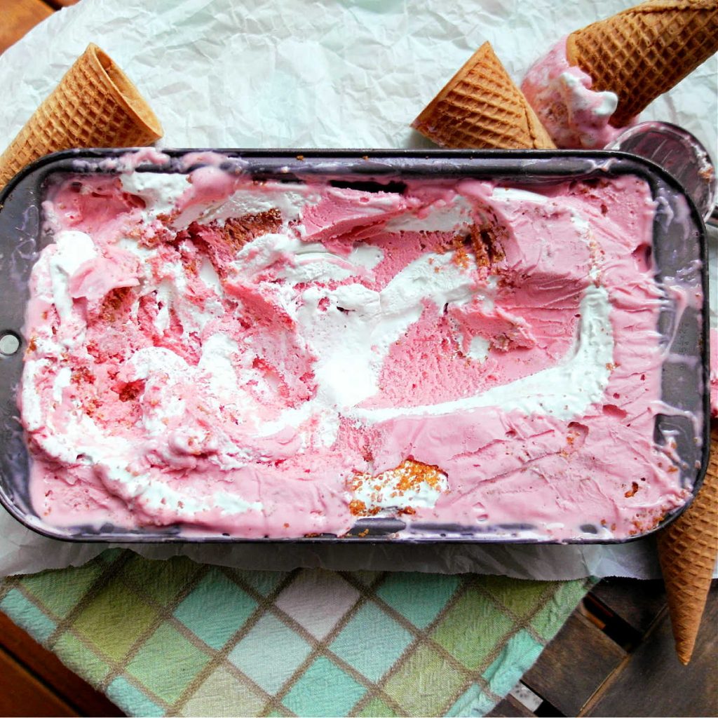A metal loaf container of strawberry cheesecake ice cream with swirls of key lime marshmallow swirl and graham cracker crumbles in it.