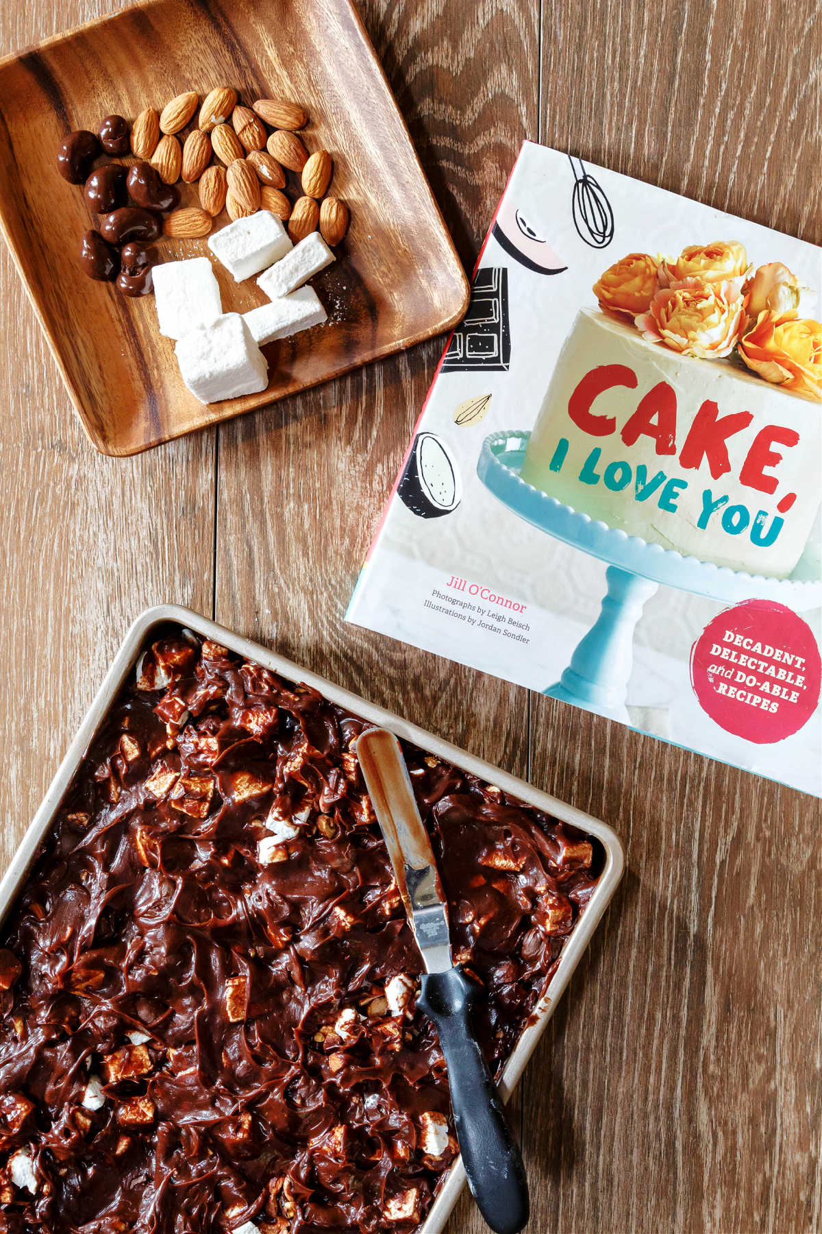Overhead shot of chocolate sheet cake with rocky road frosting, the cookbook "Cake, I Love you", and a square wooden plate with chocolate covered cashews, toasted almonds, and homemade marshmallows.