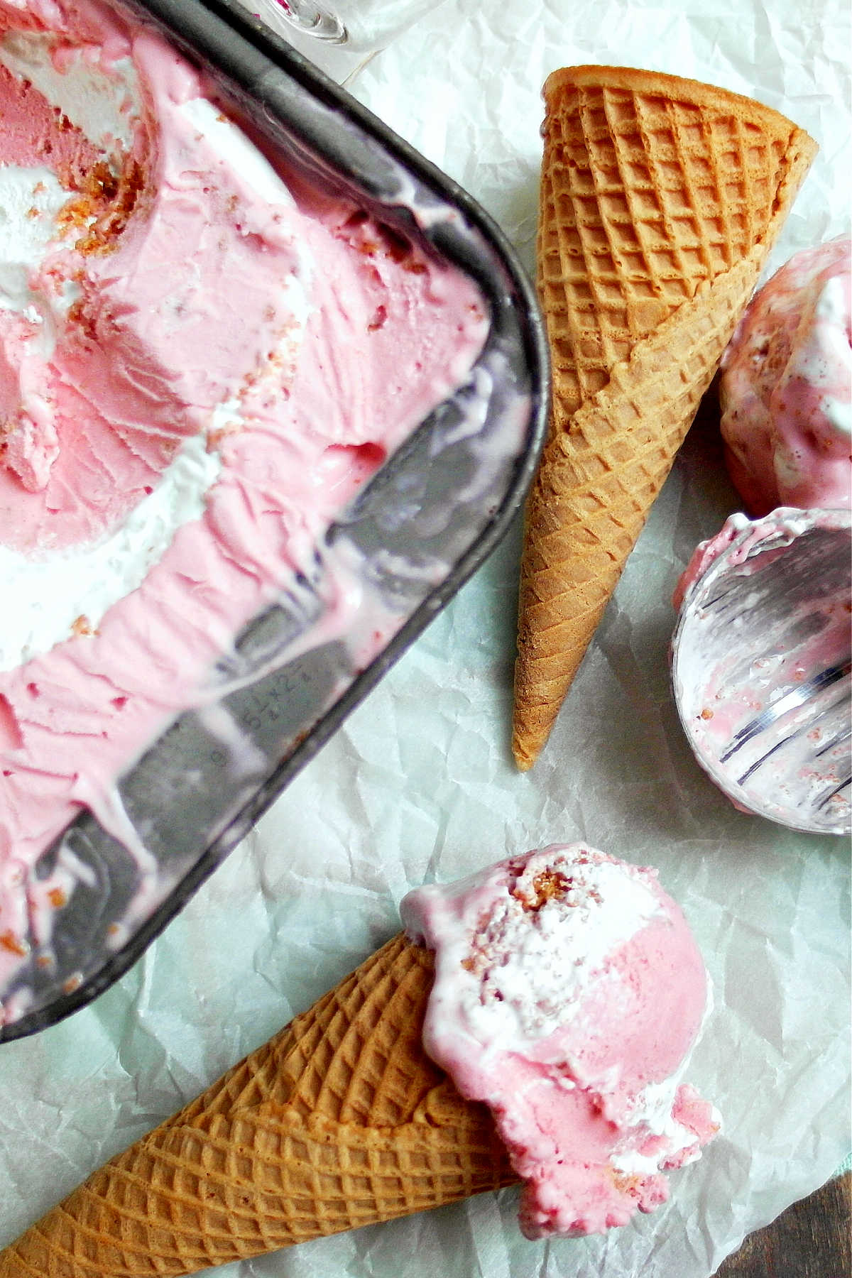 A metal pan of strawberry cheesecake ice cream, an ice cream cone and a serving of the ice cream in a cone.
