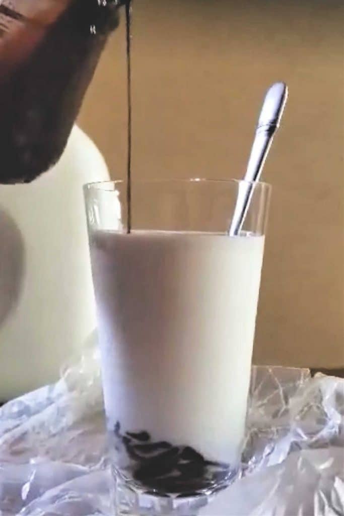 A glass of milk with chocolate syrup pouring in from the top and pooling in the bottom of the milk. A long metal spoon rests in the milk, ready for stirring.