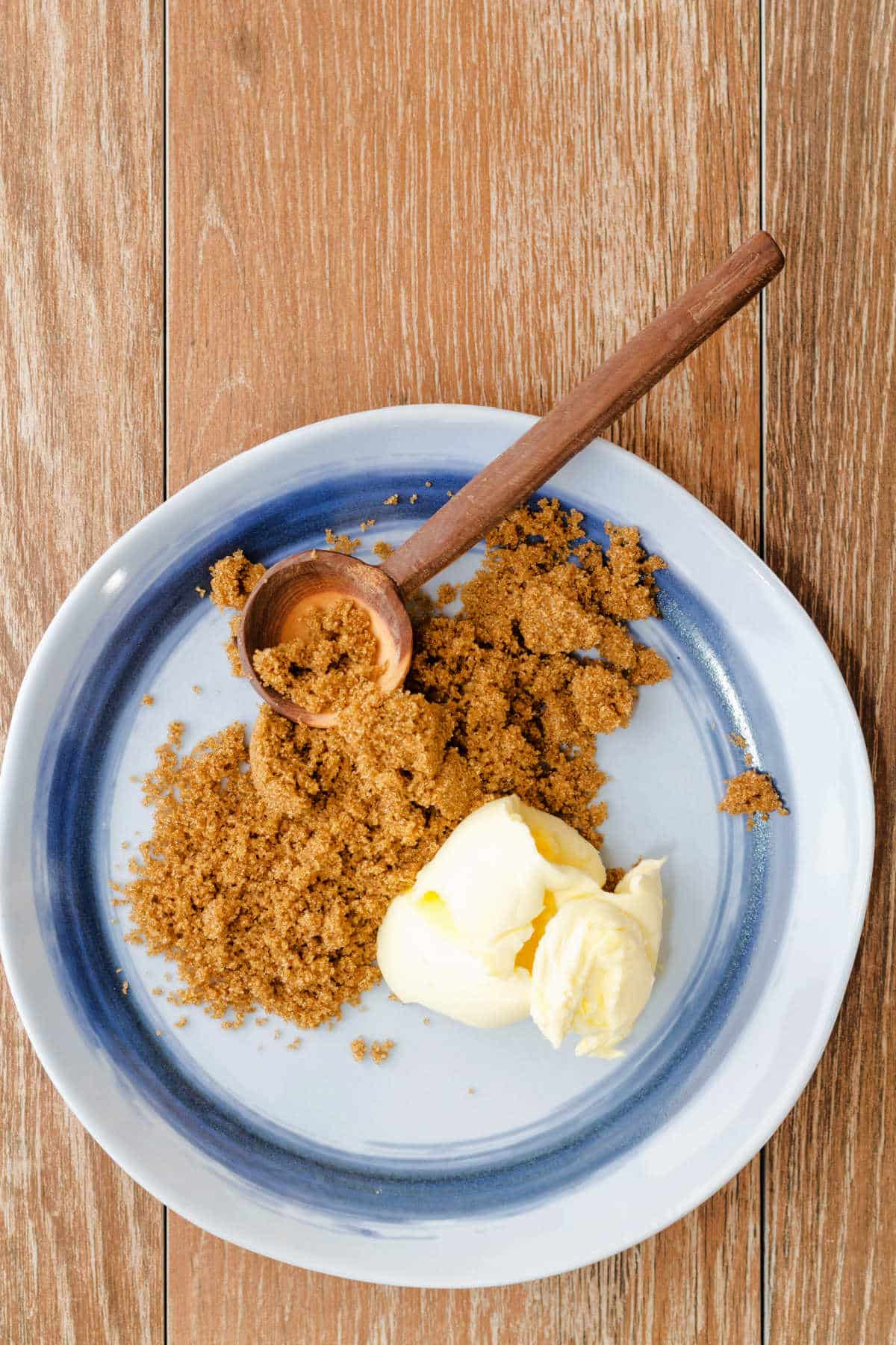 An overhead shot of a blue plate with a wooden spoon, dark brown sugar, and butter.