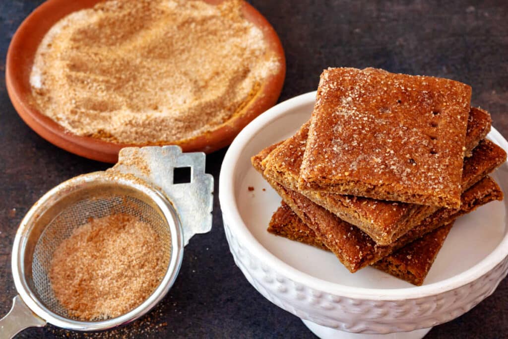 A stack of cinnamon sugar dusted Graham crackers with a small sifter and dish of cinnamon sugar.