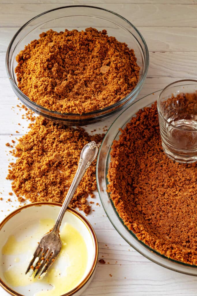 An overhead shot of a bowl of Graham cracker crumbs made from homemade Graham crackers, a dish with some melted butter in it, and a glass pie dish lined with homemade Graham cracker crust.