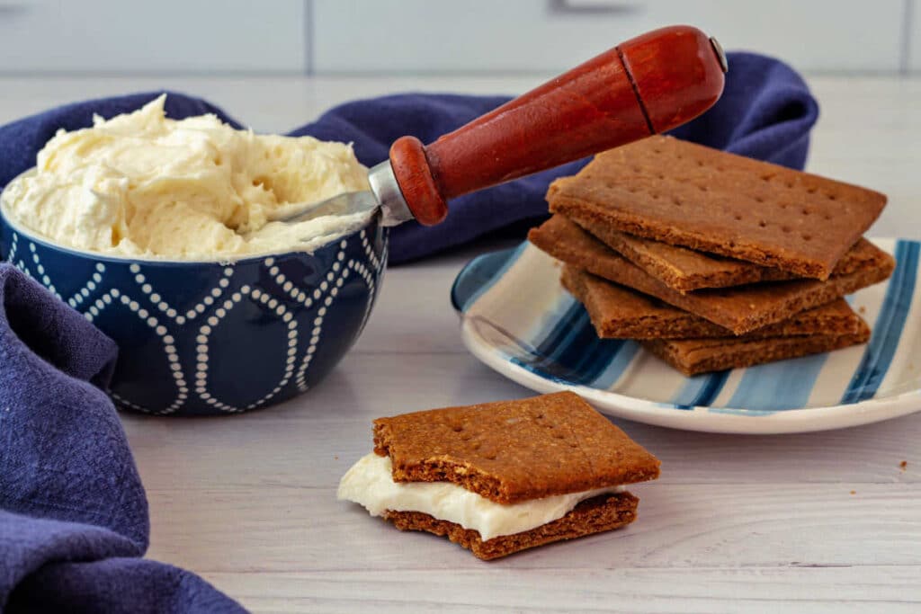 A stack of Graham crackers on a plate next to a bowl of vanilla buttercream. In front, two Graham crackers are sandwiched together with the frosting.