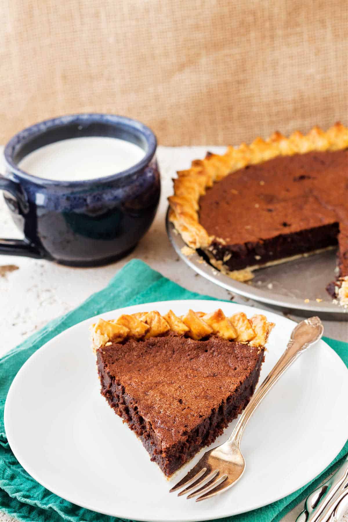A slice of chocolate pie on a white plate with the rest of the pie in the background and a mug of cold milk.