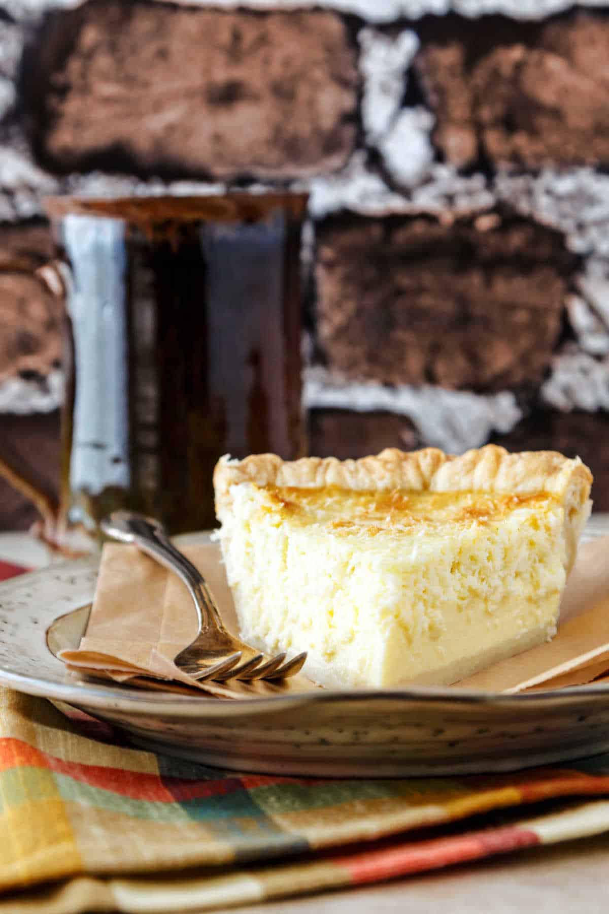 A slice of coconut custard pie on a beige plate with a brown mug to the left and a brick background.