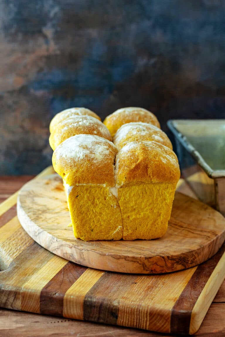 A loaf of pumpkin bread on a stack of cutting boards. The loaf is made of 6 balls of dough all baked together in a loaf pan, and the top is dusted with flour.