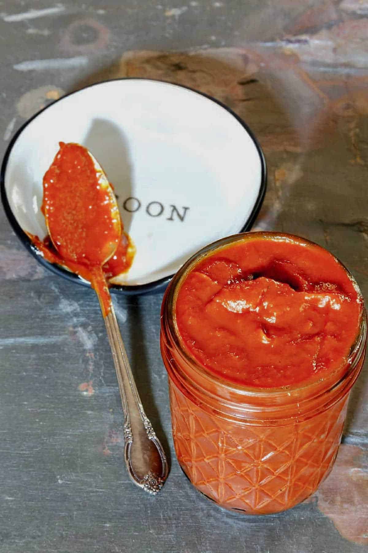 A glass canning jar of spicy ketchup with some ketchup on a spoon on a spoon rest.