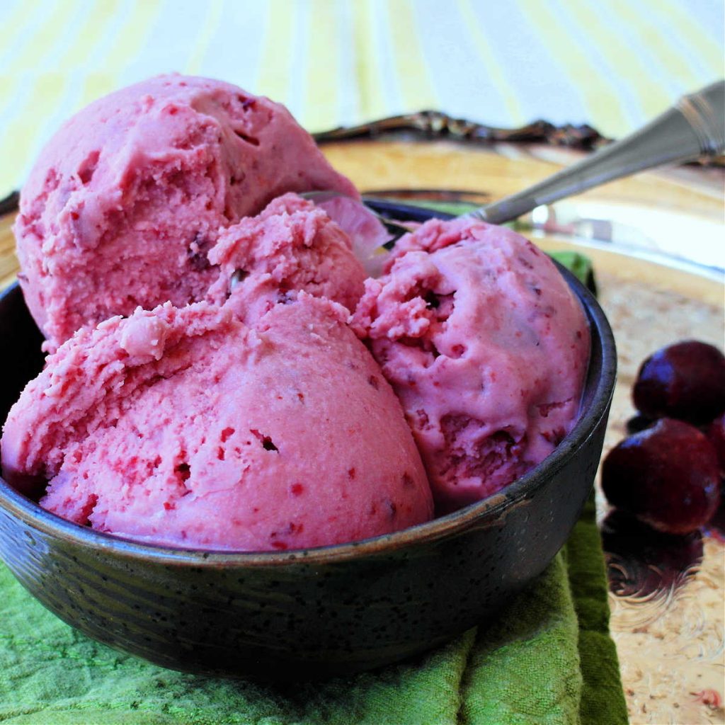 A bowl of pink sour cheery ice cream with a metal spoon. Fresh cherries are on the side.
