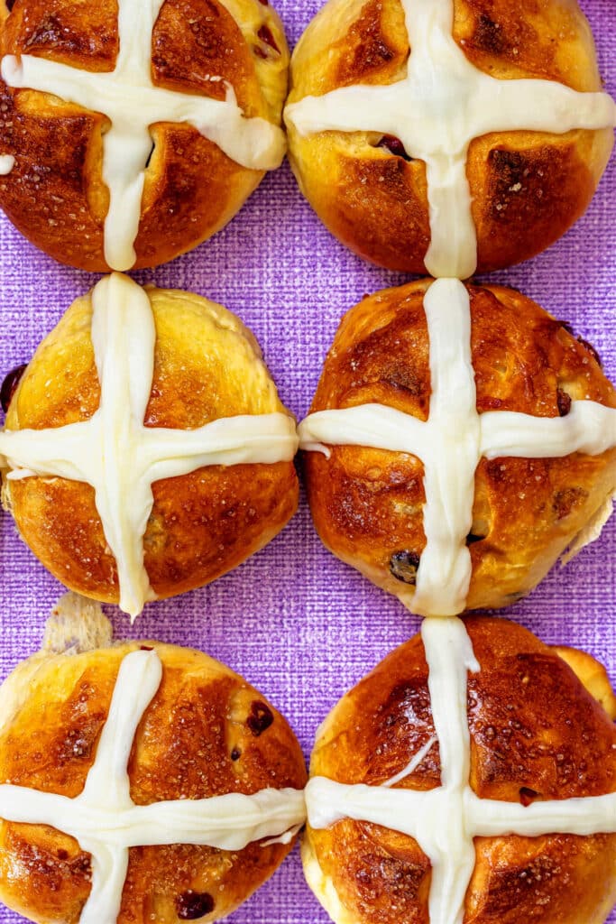 An overhead shot of six hot cross buns all lined up with their icing crosses touching on a purple linen surface.