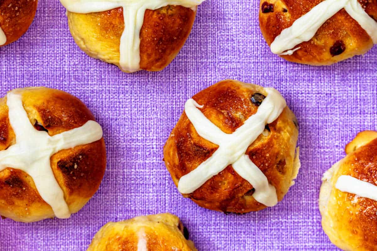 An overhead shot of hot cross buns with icing crosses on them arranged on a lavender linen surface.