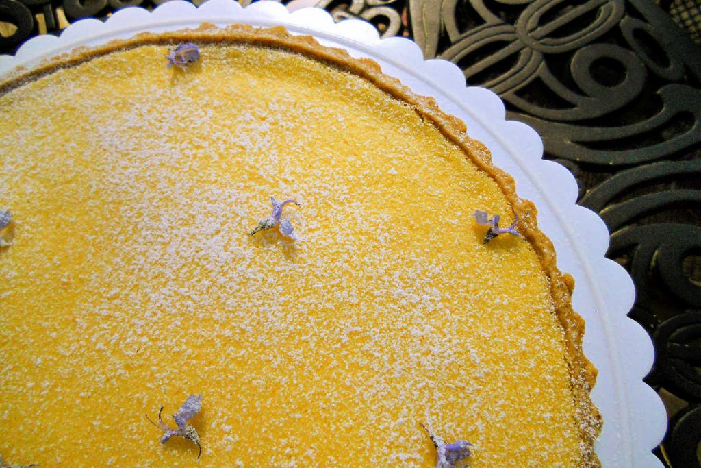 Overhead partial shot of a lemon tart decorated with powdered sugar and small purple rosemary blossoms.