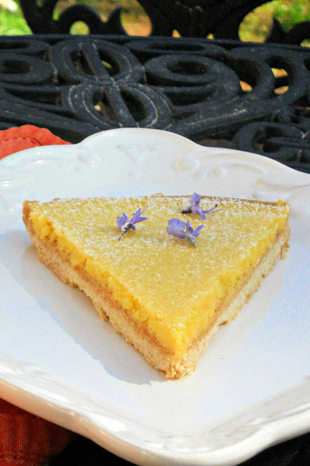 A slice of lemon tart on a white plate. The tart is decorated with small purple rosemary flowers.