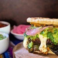 Close up of the side of a burger with pupusa buns and toppings dripping down the sides.