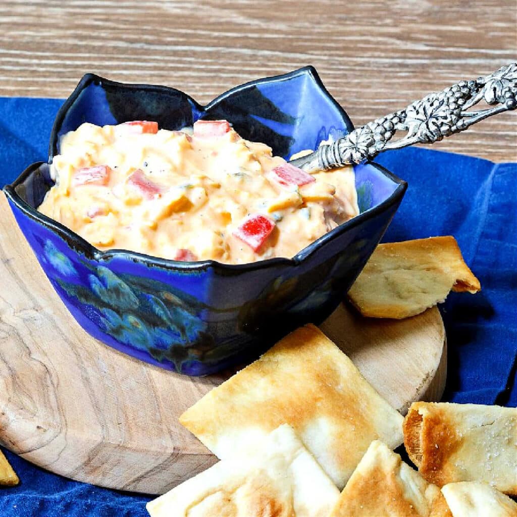 A square image of a blue, fluted bowl or pimento cheese spread on a small cutting board with a blue napkin underneath. There are some square crackers in the foreground and a small, silver metal spreader is in the dish of pimento cheese.