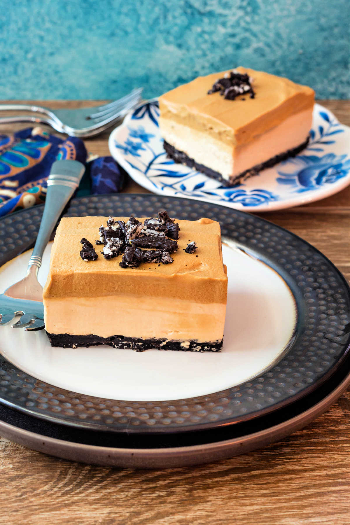 A vertical image with a square slice of dessert with chocolate crust, a tan cheesecake layer, and a darker tan whipped coffee layer topped with crushed chocolate cookies on a plate with a similar plated dessert in the background.