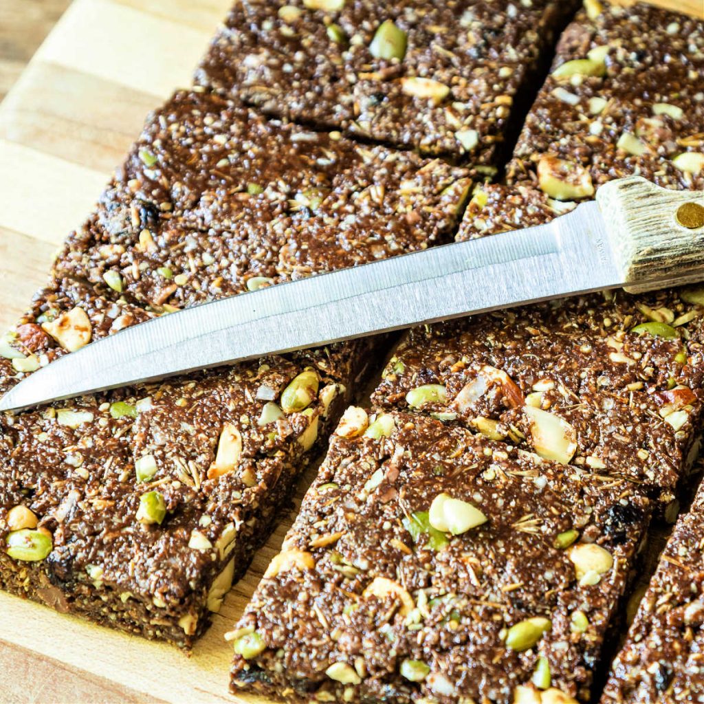 A high-angle shot of cut granola bars with pepitas in them on a cutting board with a wooden-handled knife lying on top of the cut bars.