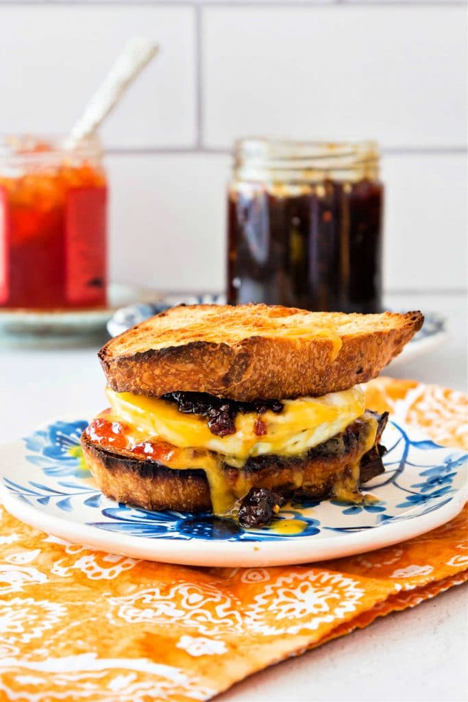 A toasted breakfast sandwich on a plate with jars of bacon jam and pepper jelly in the background.