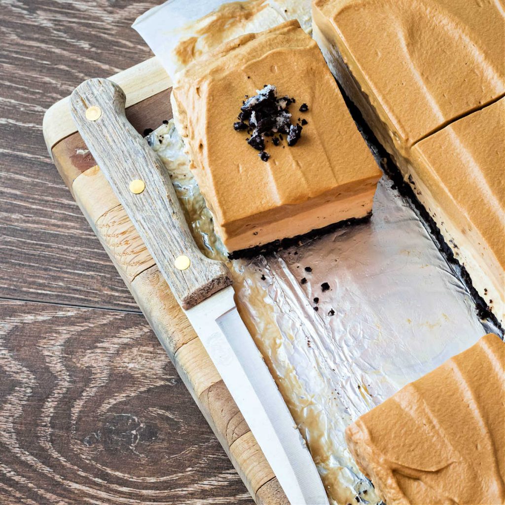 A high-angle shot of a wooden cutting board on a wooden background. On the cutting board are sliced coffee cheesecake bars, one with crushed chocolate cookies on top.