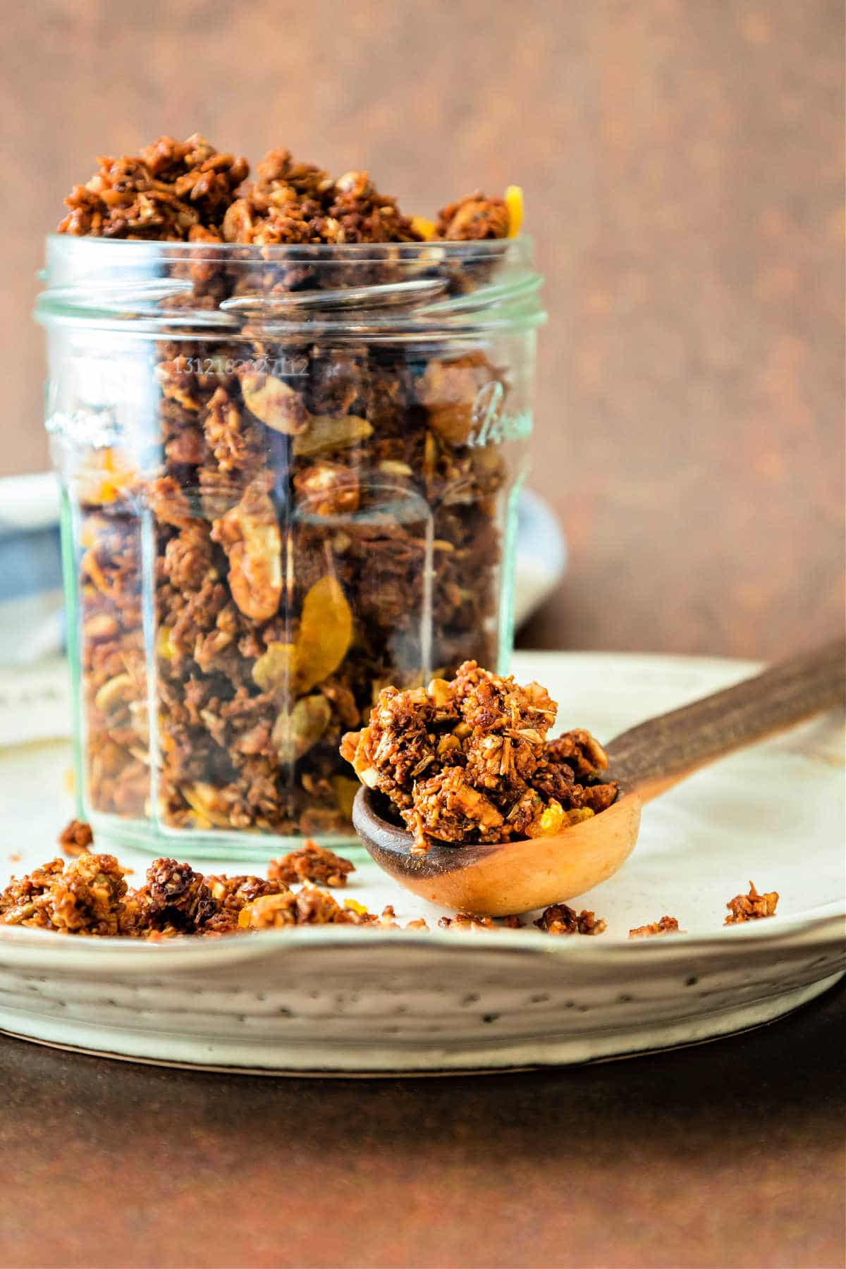 A jar of spent grain granola with chopped dried apricots in a glass jar on a beige plate.