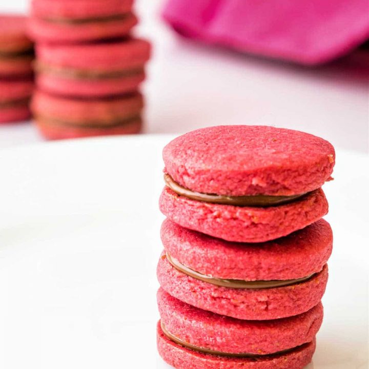 Vertical image of 3 raspberry pink sandwich cookies with chocolate filling stacked on a white plate with more stacks of cookies in the background.