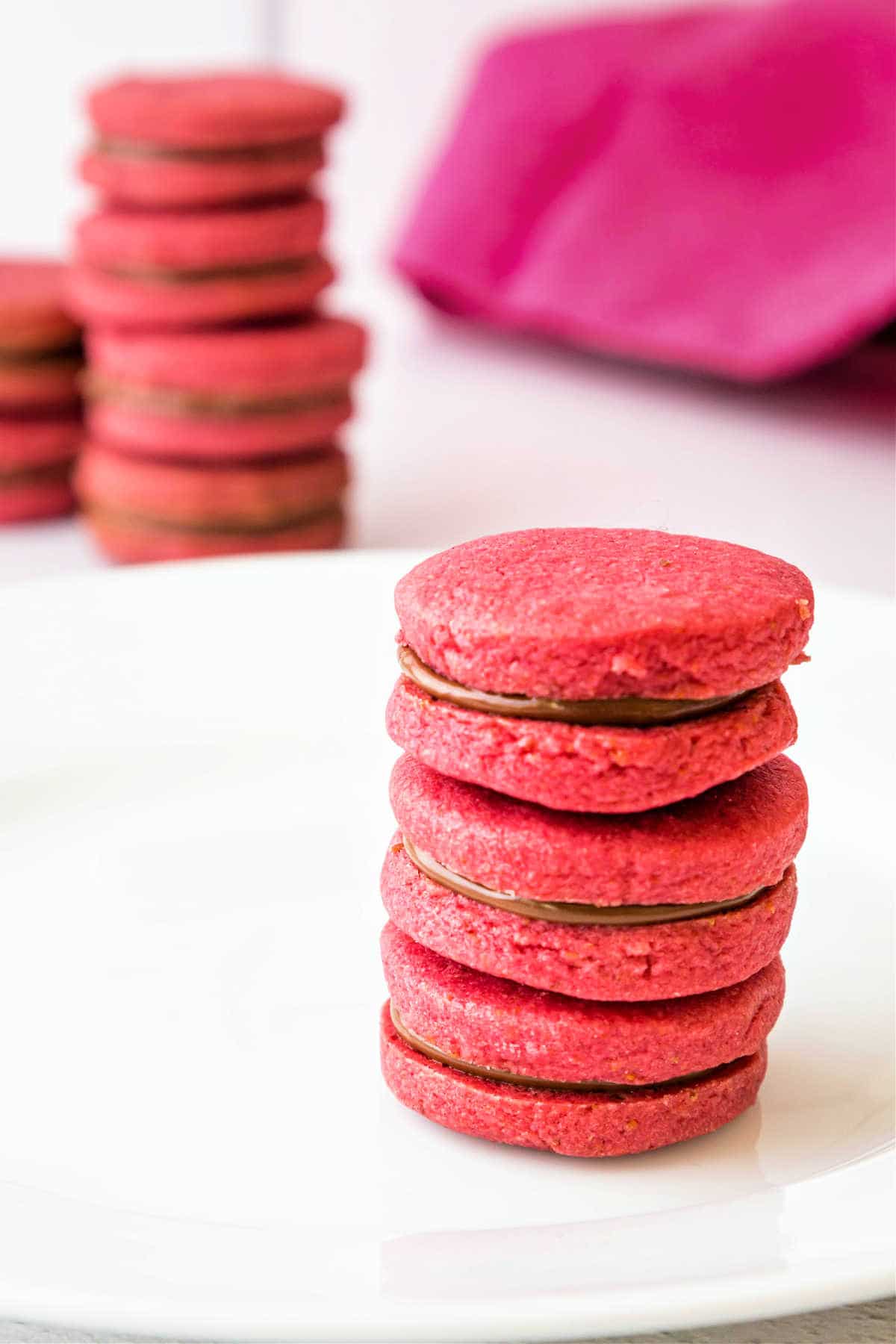 Vertical image of 3 raspberry pink sandwich cookies with chocolate filling stacked on a white plate with more stacks of cookies in the background.