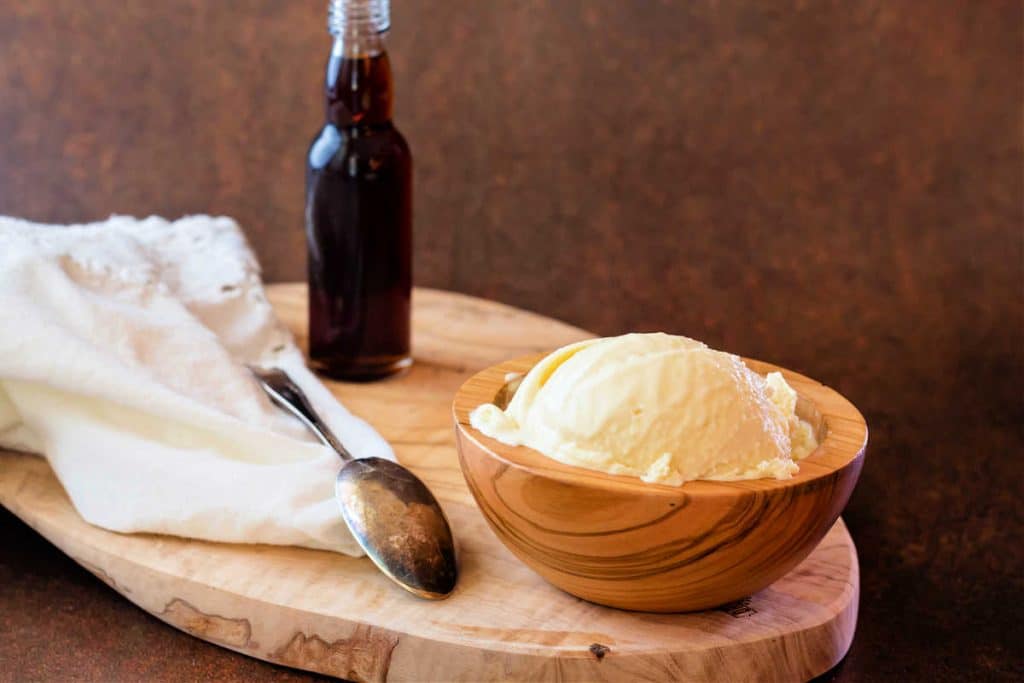 A wooden bowl of ice cream with a spoon, beige napkin, and a little bottle of vanilla extract all on an oval wooden board against a brown background.