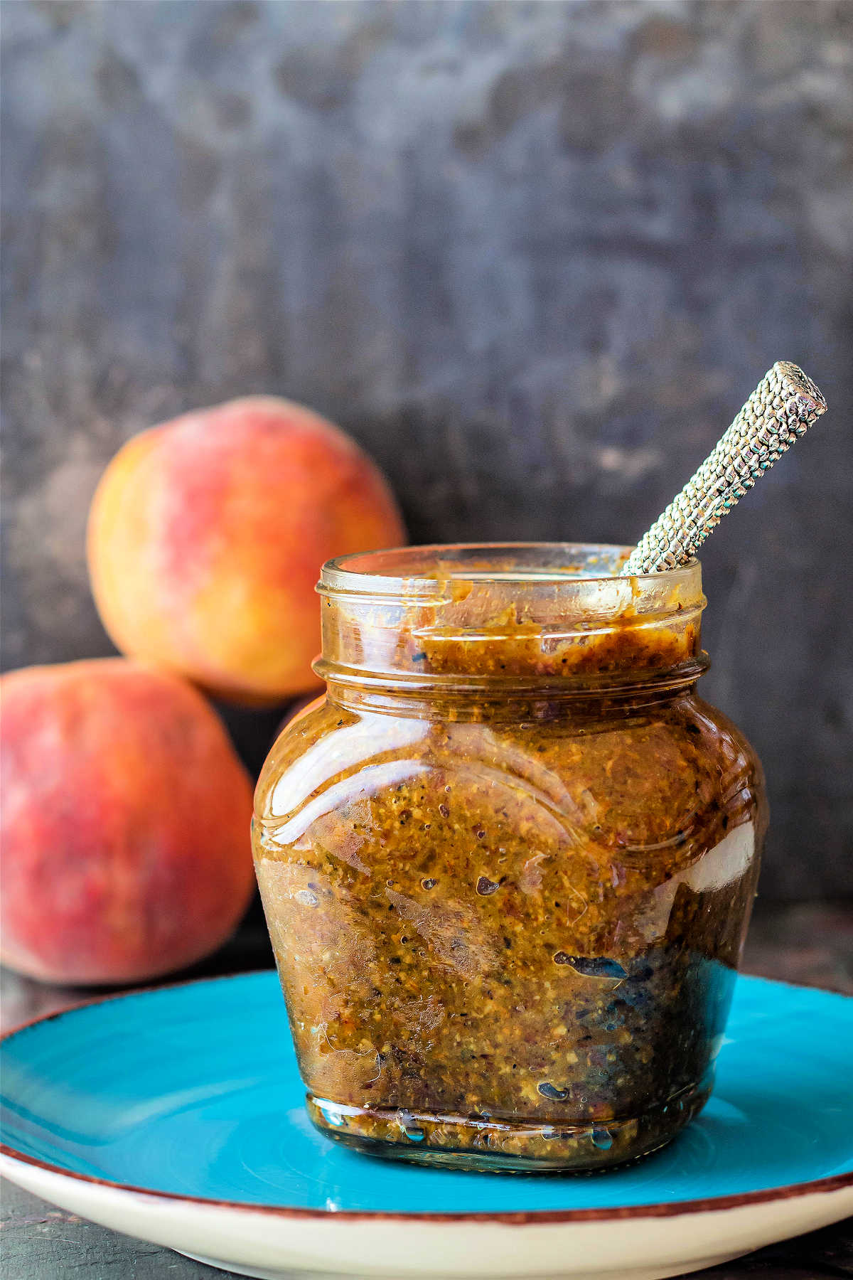 A glass jar of peach bacon jam with 3 peaches in the background.
