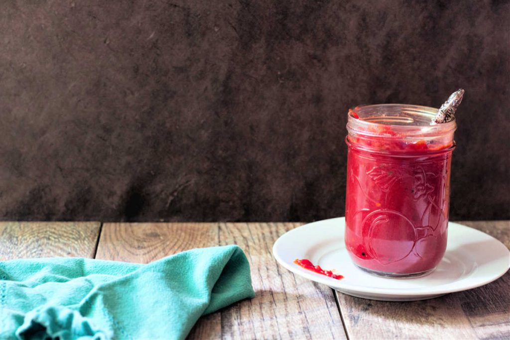 A jar of red jam on a white plate with a turquoise napkin all shot against a dark background.