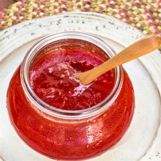A glass jar of bright red-orange jam with a wooden spoon in it.