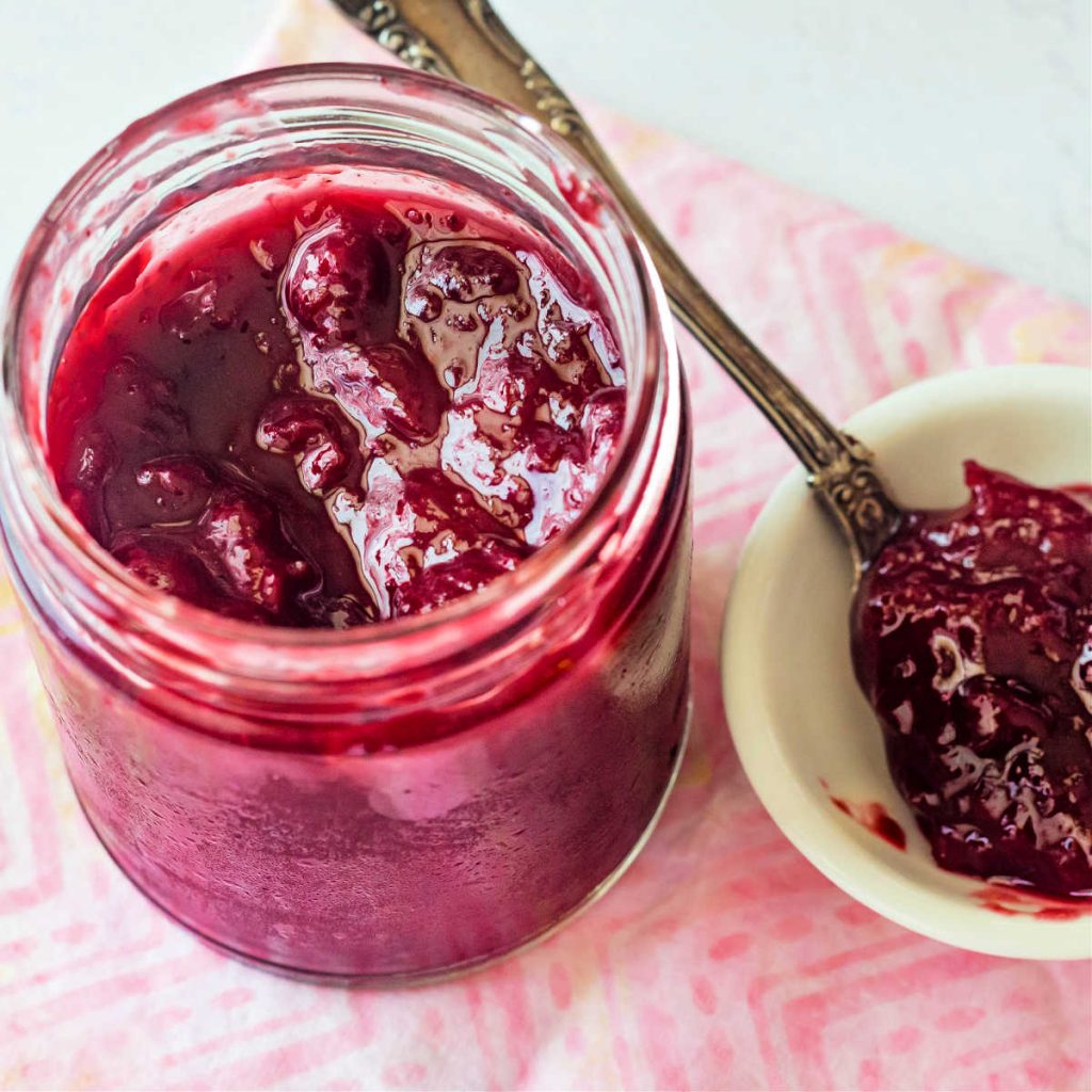 A shot into the top of an open jar of savory plum jam with a spoonful of jam in a small white dish next to it.