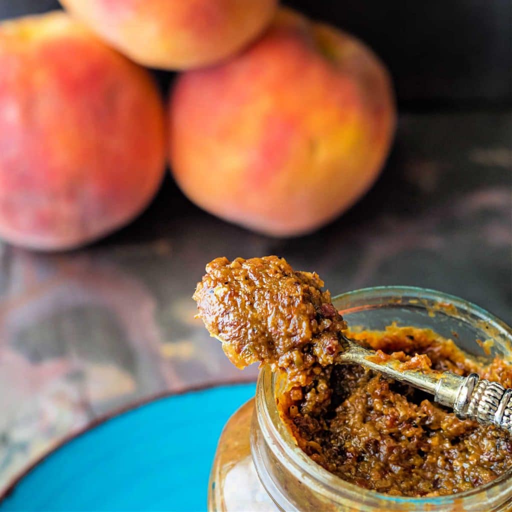A square image with a jar of bacon peach jam partially in the frame with a spoonful of the jam balanced on top. In the top of the frame, there are 3 whole peaches, slightly out of focus.