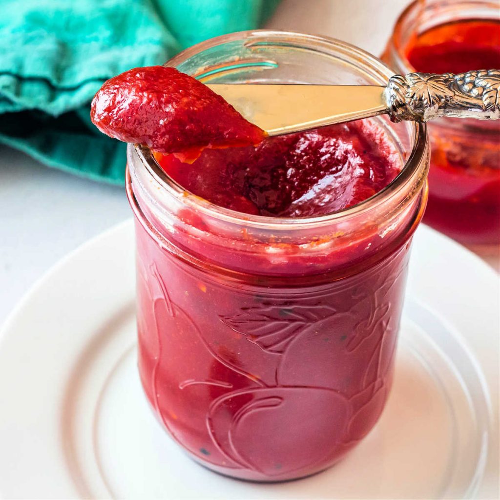 A close up of a jar of bright red tomato jam on a white plate with a silver knife.