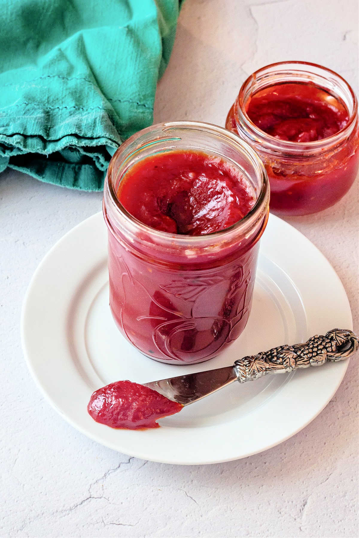 Two jars of tomato jam, one on a white plate with a knife with tomato jam on it. There's a teal napkin in the background.