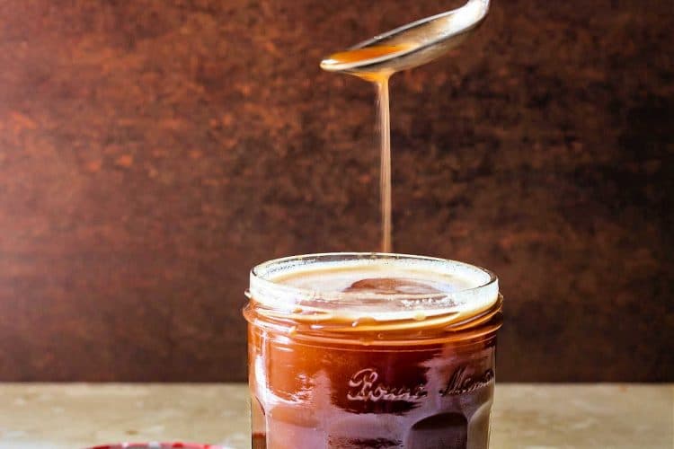 An open and full jar of butterscotch syrup with the lid next to it. There's a hand holding a spoon with syrup dripping down into the open jar.