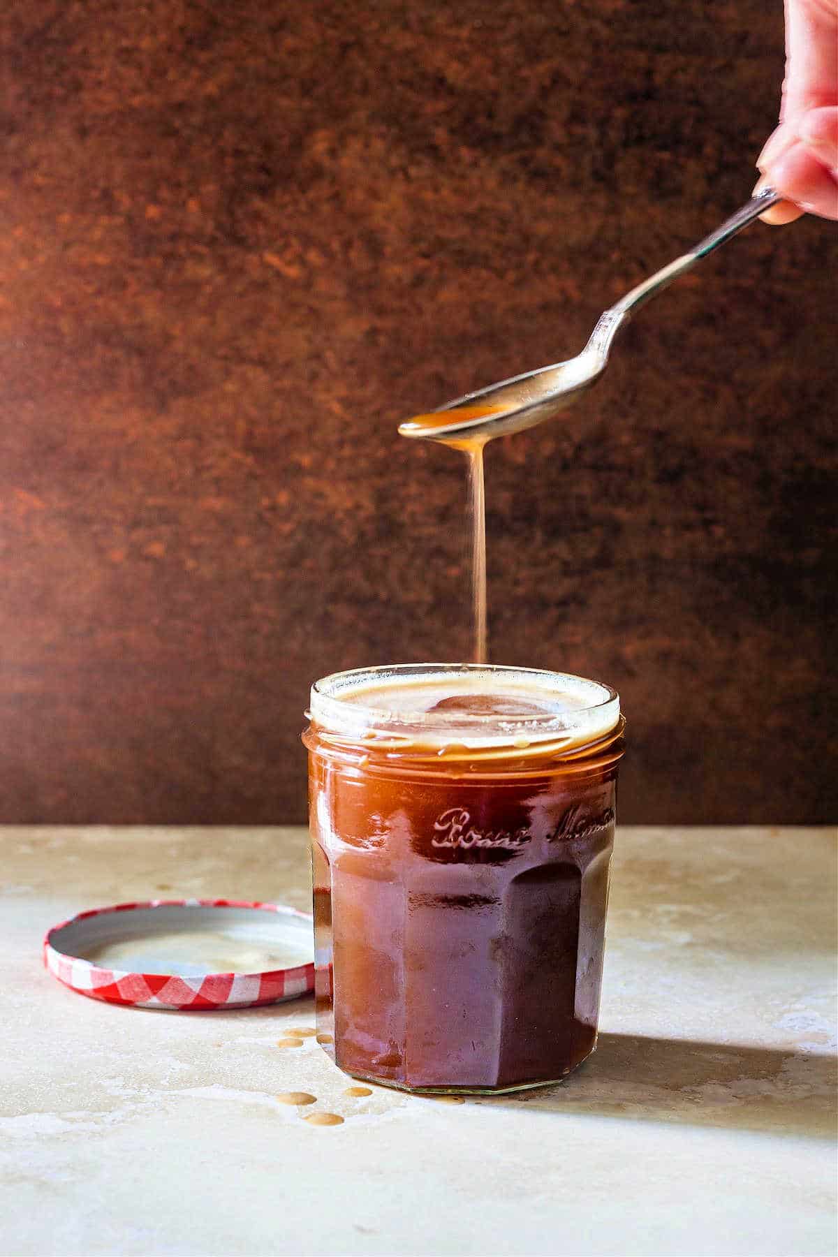 An open and full jar of butterscotch syrup with the lid next to it. There's a hand holding a spoon with syrup dripping down into the open jar.