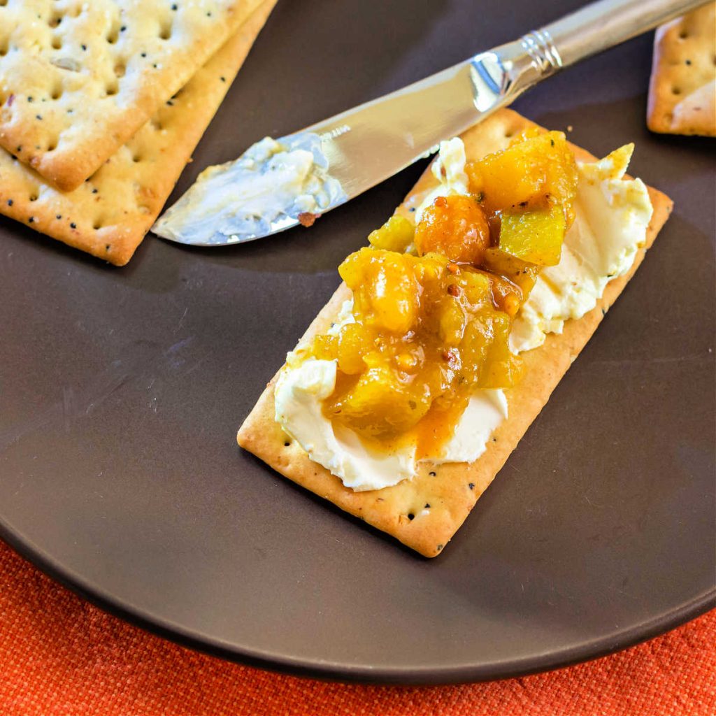 A rectangular cracker spread with cream cheese and topped with apple green tomato chutney on a brown plate.