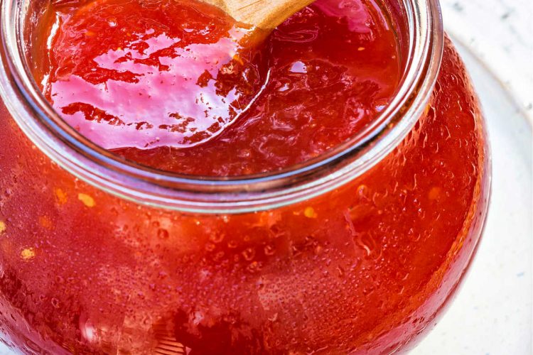 A vertical shot of a jar of peach tomato jam with a small wooden spoon in it.