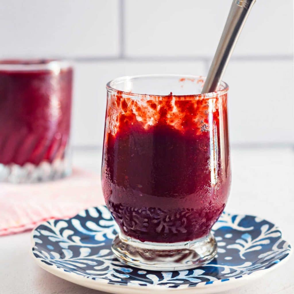 A clear glass jar full of deep reddish purple jam on a blue patterned plate with a knife in the jar.