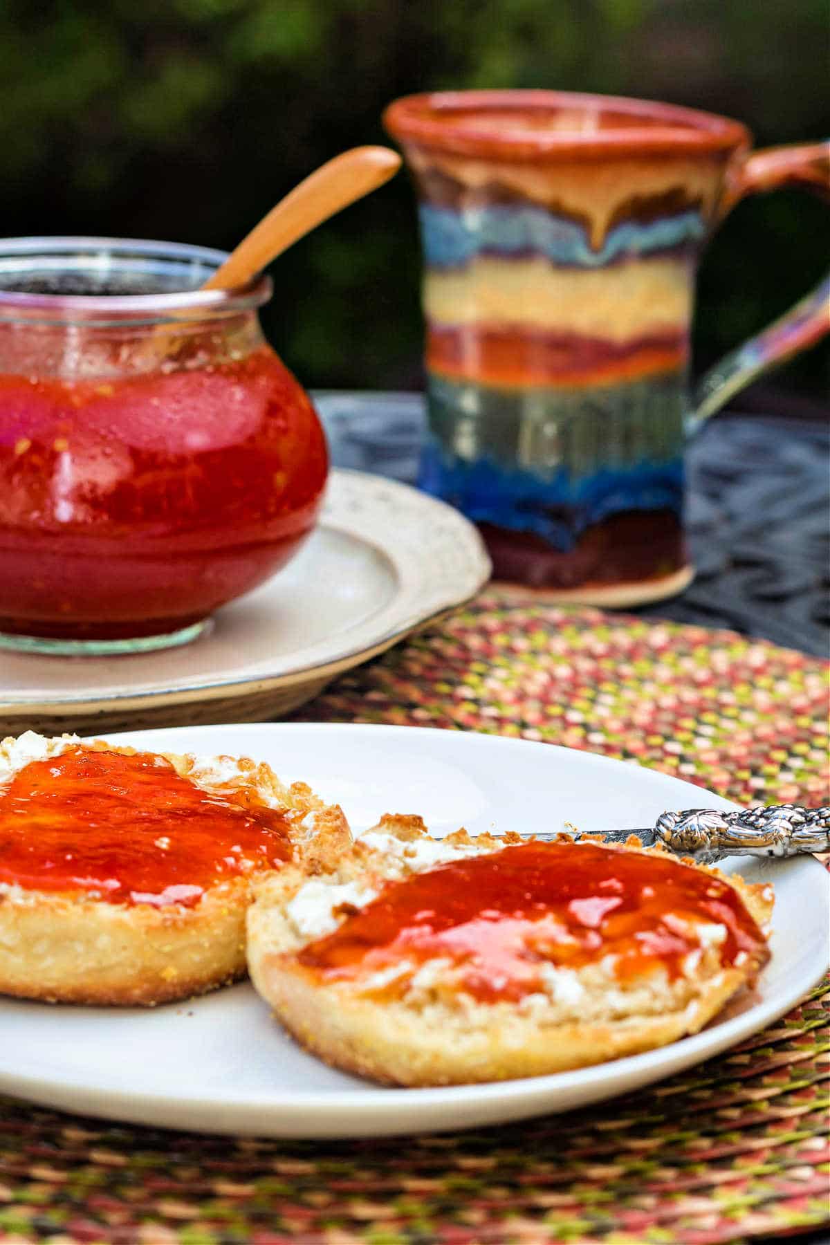 A white plate with two pieces of English muffin on it spread with jam. The jar of jam is in the background along with a multi-colored mug.