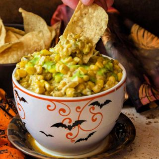 A bowl of deviled corn dotted with green salsa with a hand reaching a tortilla chip into it.