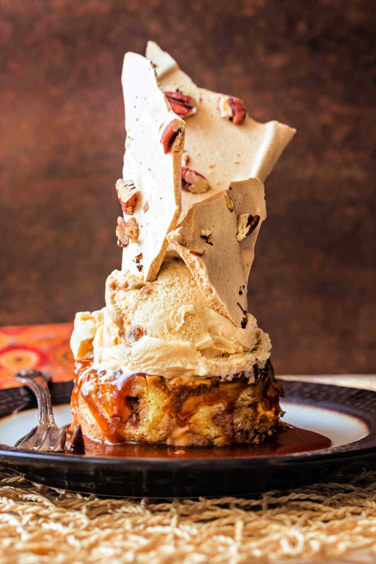 A dramatic shot of a piece of bread pudding in a pool of caramel topped with caramel ice cream with tall pieces of crispy meringue stuck in the ice cream.
