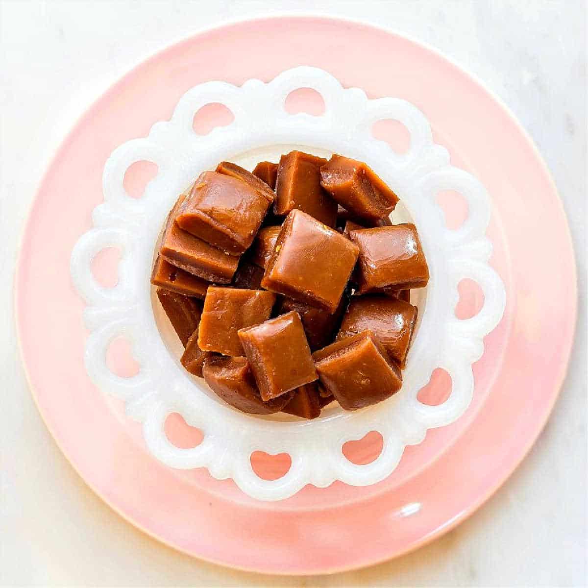 An overhead shot of a white candy dish on a pink plate filled with squares of dark caramel candy.
