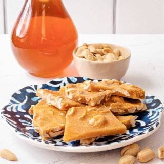Pieces of peanut brittle on a blue plate with a small dish of peanuts and a jar of honey in the background.